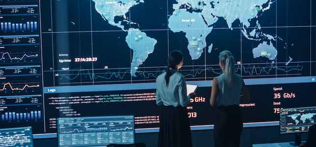 Two Software Engineers Talking Next to a Big Screen in a Modern Monitoring Office with Live Analysis Feed with Charts.